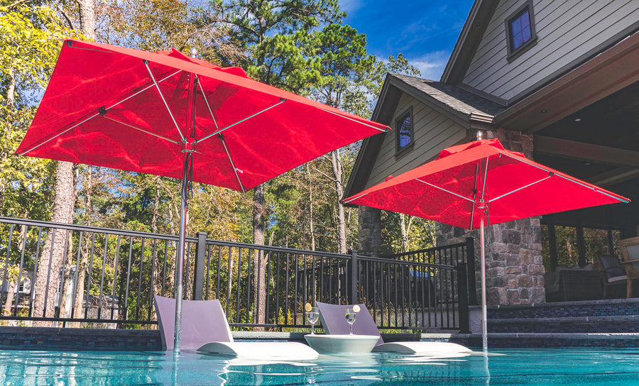 Two Ledge Lounger white lounge chairs with attached drink tables are partially submerged in a pool, shaded by two Premier Aluminum Umbrella - 13 Octagon Pulley models. The pool, lined with stone and surrounded by trees, is next to a stone exterior house.