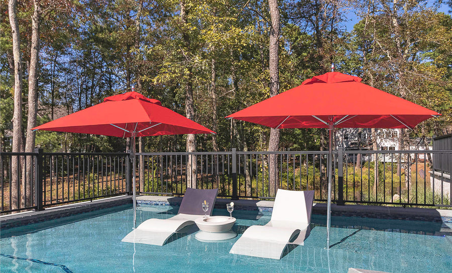 The poolside scene features two white lounge chairs partially submerged in water, each with a glass of sparkling beverage. Two Ledge Lounger Premier Aluminum Umbrellas - 11 Octagon Pulley provide shade with wind-resistant, marine-grade fabrics. A black metal fence and trees complete the tranquil backdrop.