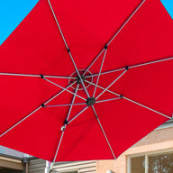 The Ultra Cantilever Umbrella - 10 Square by Ledge Lounger opens a vibrant red canopy above a patio, set against blue skies and modern architecture. Rechargeable lights subtly illuminate the area, highlighting large windows and a sleek, sloped roof in contemporary style.