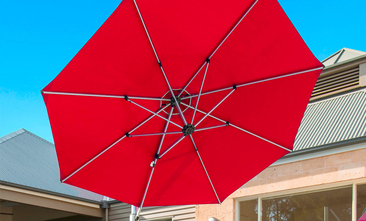The Ultra Cantilever Umbrella - 10 Square by Ledge Lounger opens a vibrant red canopy above a patio, set against blue skies and modern architecture. Rechargeable lights subtly illuminate the area, highlighting large windows and a sleek, sloped roof in contemporary style.