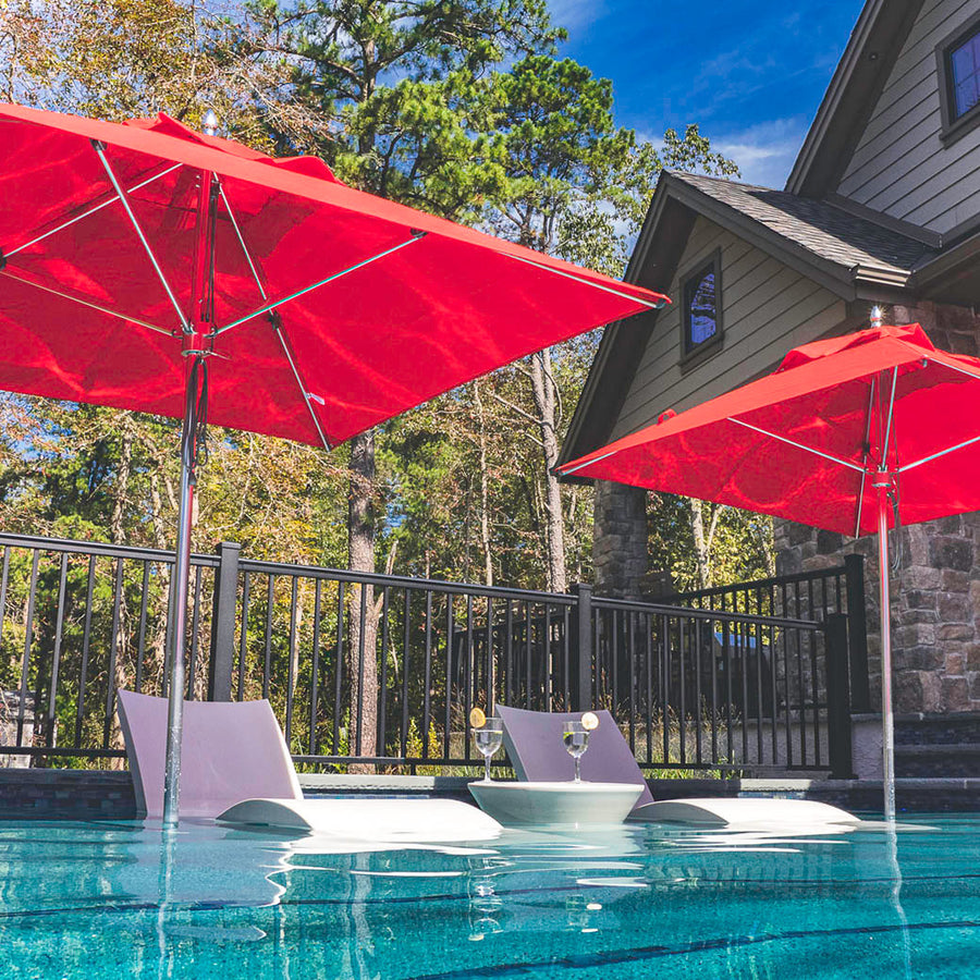 Two white lounge chairs with attached tables sit in a clear pool under the shade of two 7.5 Premier Aluminum Umbrellas with marine-grade fabric by Ledge Lounger. A stone house with green siding and black railing is surrounded by trees, all under a clear blue sky.
