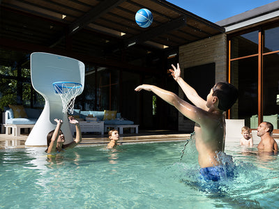 A boy in blue swim trunks jumps to shoot a basketball toward a poolside hoop. Three other children watch from the water, and two adults are in the background. The setting is an outdoor pool area with a modern house.