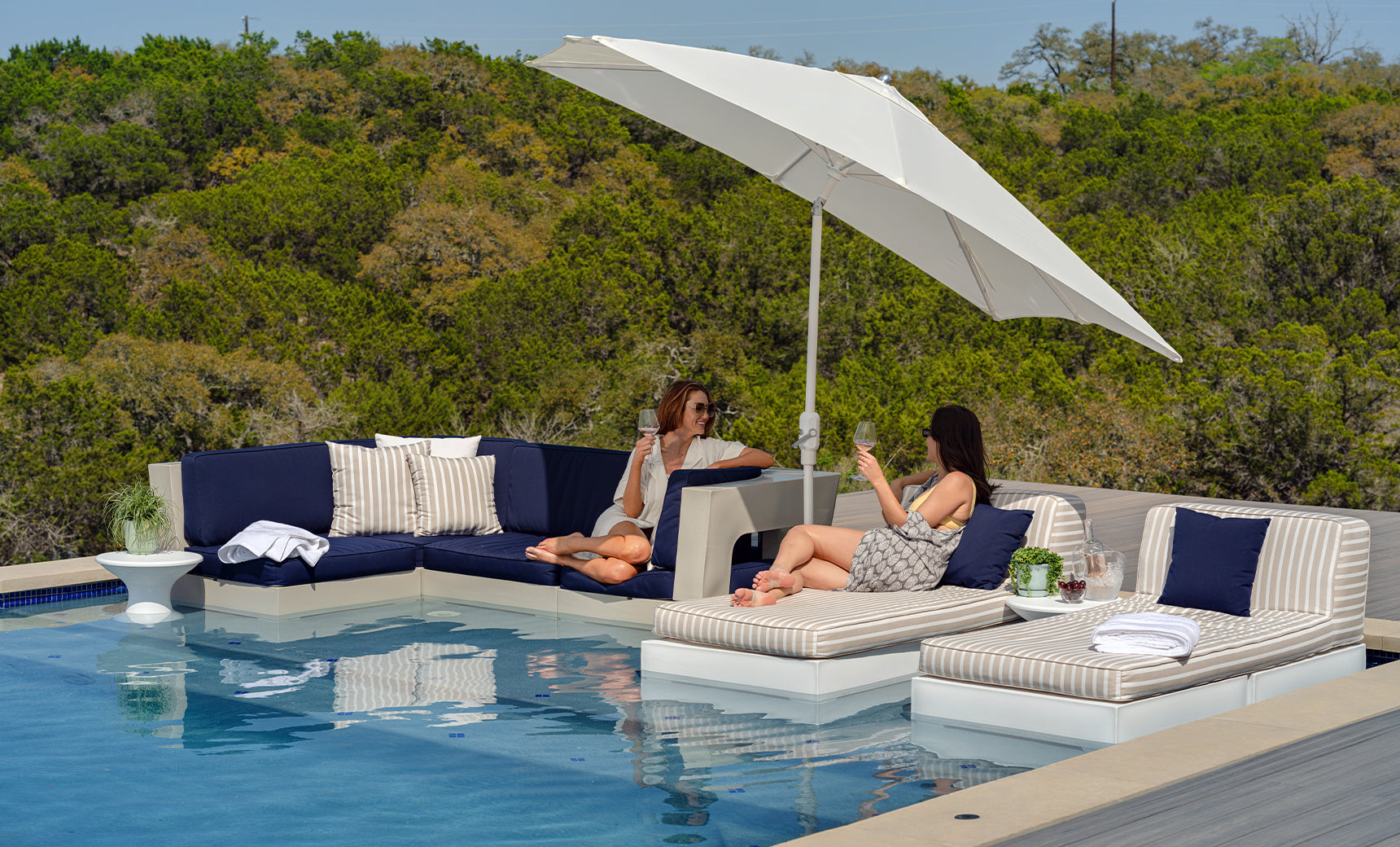 Two women relax on a luxurious patio set by the pool, enjoying resort-level luxury. They sit beneath a Ledge Lounger 7.5 Octagon Signature 360° Tilt Umbrella, surrounded by trees. The area features a navy couch and striped loungers, with a towel and drinks enhancing the serene scene.