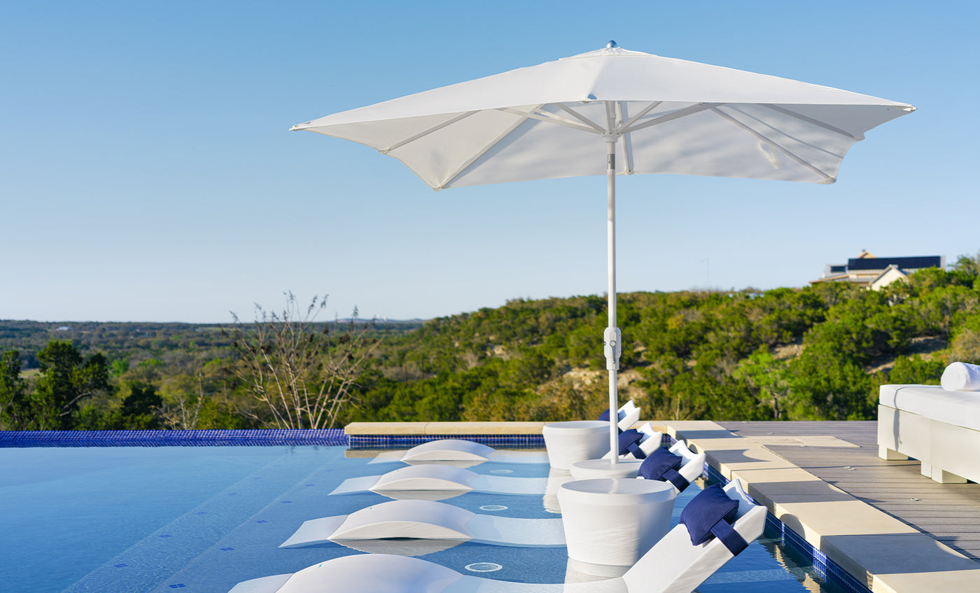 An infinity pool showcases partially submerged chairs under Ledge Loungers 7.5 Octagon Signature 360° Tilt Umbrella, offering resort-level luxury as it overlooks lush greenery and a clear sky. A white couch on the deck adds to the opulent outdoor ambiance.