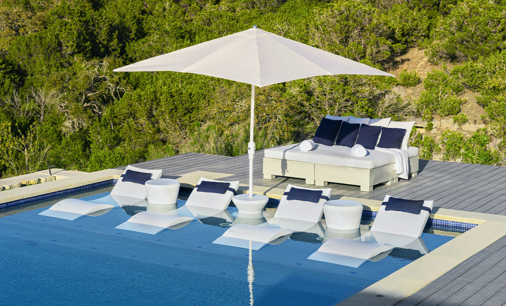 A modern poolside oasis includes white lounge chairs partially submerged in water, perfectly shaded by a Ledge Lounger 7.5 Octagon Signature 360° Tilt Umbrella. A double daybed with blue and white cushions sits on a wooden deck amidst lush greenery.