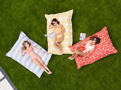 Three people in swimsuits lounge on patterned cushions on grass, each holding a magazine. One cushion is blue, one yellow with circles, and one red with a geometric design. A small speaker is placed nearby.