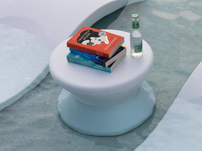 A white, round table stands in shallow water, holding three books and a bottle of Perrier. The books include Crazy Rich Asians, Big Little Lies, and Grocery. The scene suggests a relaxing poolside or beach atmosphere.
