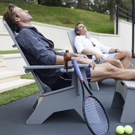 Two people relax on the Mainstay Ultimate Adirondack lounge chairs by Ledge Lounger under a clear sky. One holds a bottled drink, while a tennis racket and balls lie nearby, enjoying the serene surroundings and stylish poolside furniture.