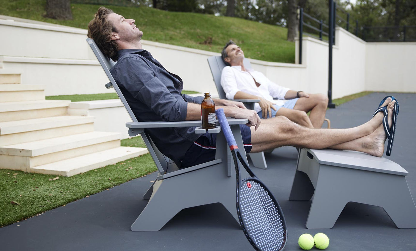 Two people relax on the Mainstay Ultimate Adirondack lounge chairs by Ledge Lounger under a clear sky. One holds a bottled drink, while a tennis racket and balls lie nearby, enjoying the serene surroundings and stylish poolside furniture.
