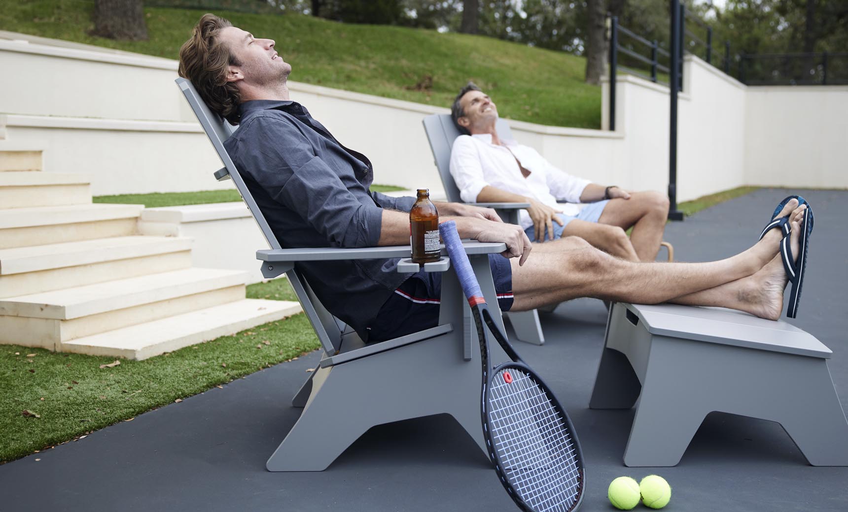 Two people relax on the Mainstay Ultimate Adirondack lounge chairs by Ledge Lounger under a clear sky. One holds a bottled drink, while a tennis racket and balls lie nearby, enjoying the serene surroundings and stylish poolside furniture.