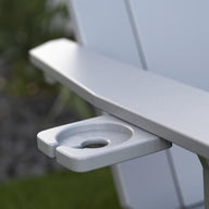 Close-up of the Mainstay Ultimate Adirondack by Ledge Lounger, a light gray outdoor lounge chair with a built-in cup holder. The blurred background of greenery and stones creates the perfect poolside relaxation setting.