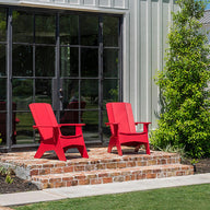 Two Mainstay Ultimate Adirondack chairs by Ledge Lounger enhance a small brick patio in front of a modern barn-style building with large windows. Surrounded by green shrubs and lawn, the scene invites relaxation outdoors under a clear blue sky.