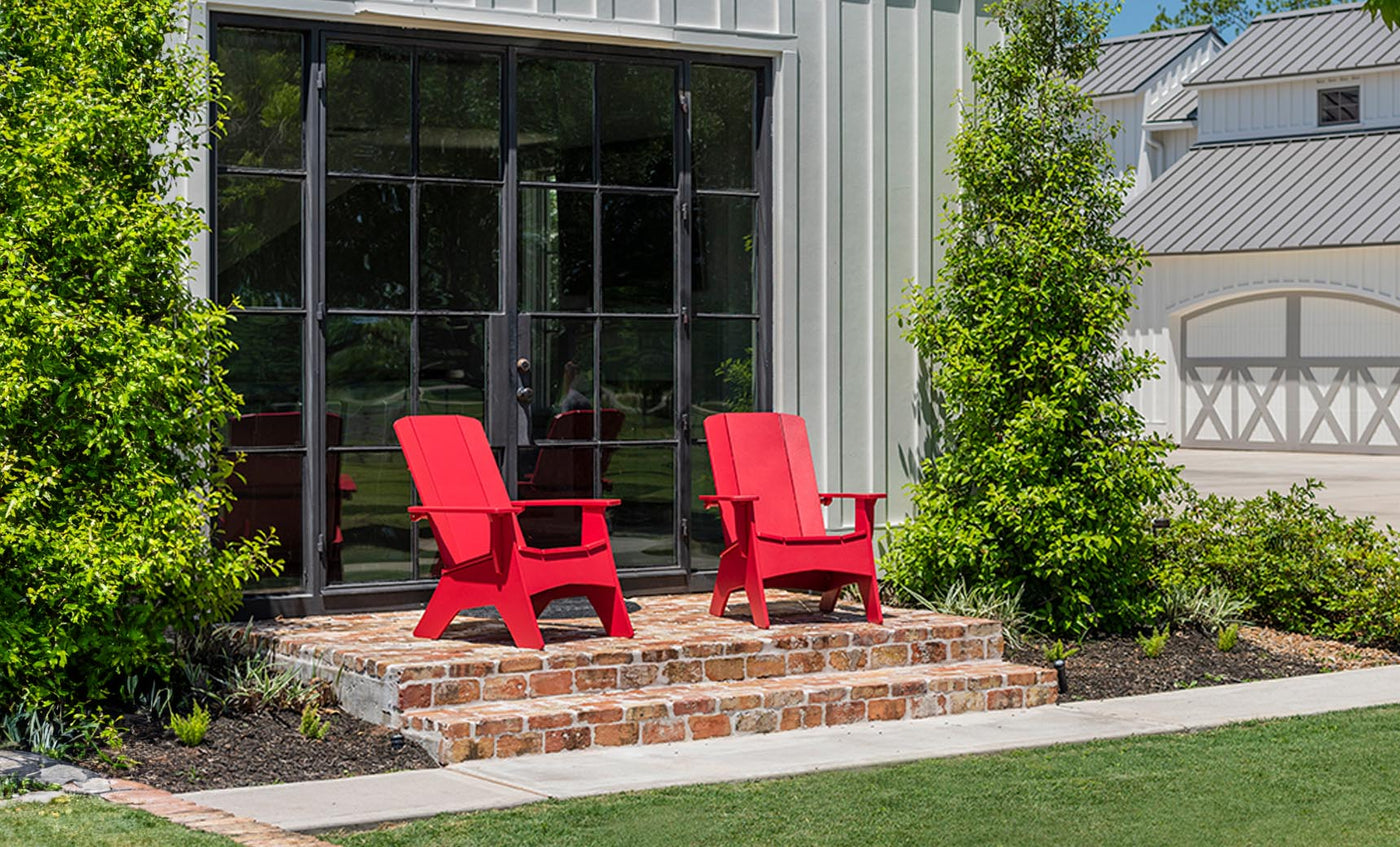 Two Mainstay Ultimate Adirondack chairs by Ledge Lounger enhance a small brick patio in front of a modern barn-style building with large windows. Surrounded by green shrubs and lawn, the scene invites relaxation outdoors under a clear blue sky.