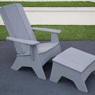 A Ledge Lounger Mainstay Adirondack Ottoman and matching chair sit on the dark patio. In the background, lush green grass borders a short white wall, creating a serene setting with high-quality materials.