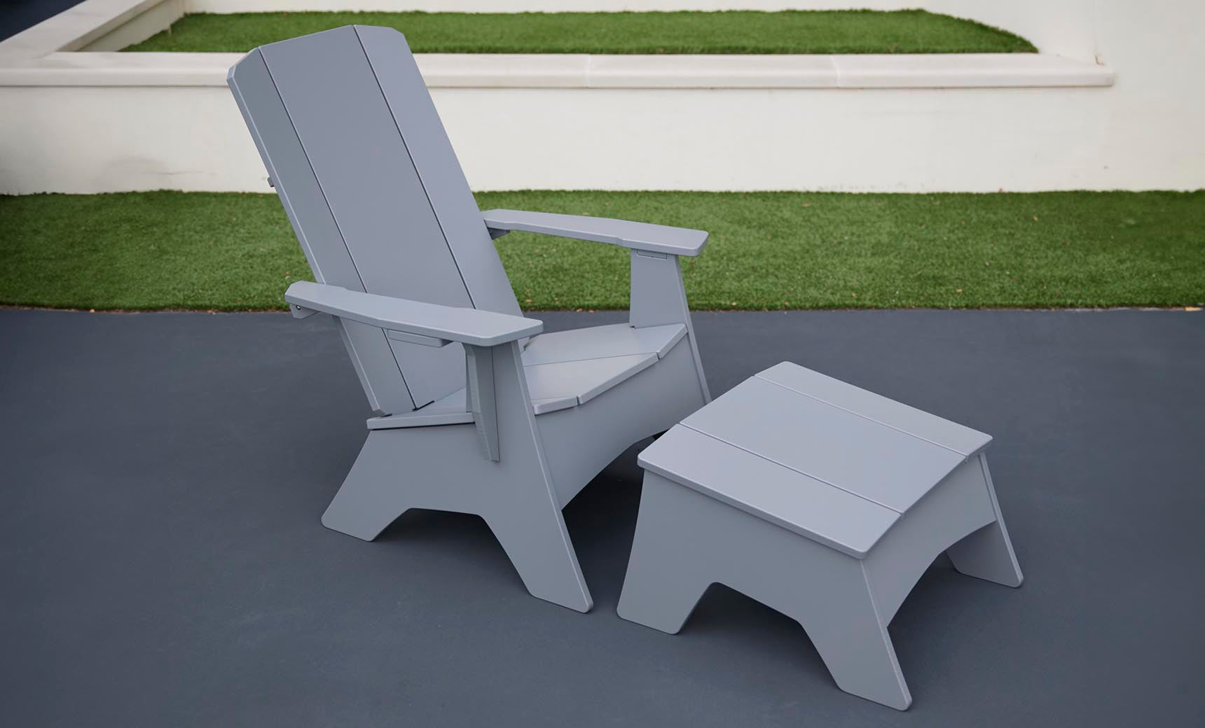 A Ledge Lounger Mainstay Adirondack Ottoman and matching chair sit on the dark patio. In the background, lush green grass borders a short white wall, creating a serene setting with high-quality materials.