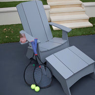 A gray Ledge Lounger chair features a matching Mainstay Adirondack Ottoman on the outdoor patio. Beside it lie two tennis rackets and balls, with a rolled-up blue towel over the arm next to a cup of pink drink, all set against steps and lush grass.