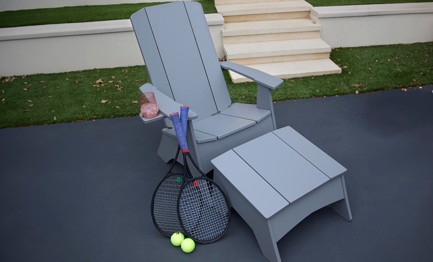 A gray Ledge Lounger chair features a matching Mainstay Adirondack Ottoman on the outdoor patio. Beside it lie two tennis rackets and balls, with a rolled-up blue towel over the arm next to a cup of pink drink, all set against steps and lush grass.