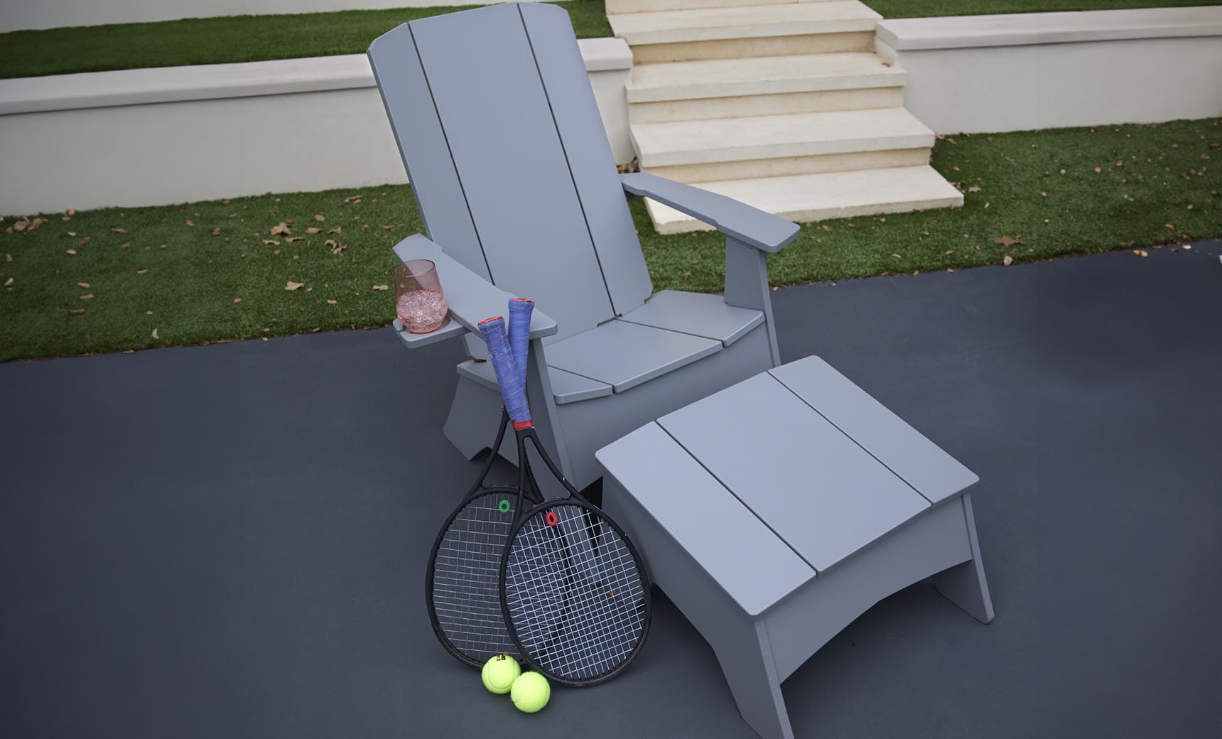 A gray Ledge Lounger chair features a matching Mainstay Adirondack Ottoman on the outdoor patio. Beside it lie two tennis rackets and balls, with a rolled-up blue towel over the arm next to a cup of pink drink, all set against steps and lush grass.