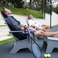 Two men relax on Ledge Lounger patio chairs with tennis rackets and balls nearby. One sips a drink as they both bask in the sun, eyes closed. Mainstay Adirondack Ottomans and steps leading to a grassy area add durability and style to the scene.