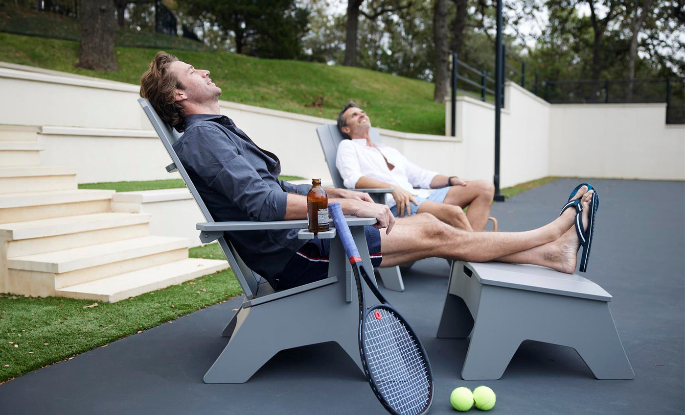 Two men relax on Ledge Lounger patio chairs with tennis rackets and balls nearby. One sips a drink as they both bask in the sun, eyes closed. Mainstay Adirondack Ottomans and steps leading to a grassy area add durability and style to the scene.