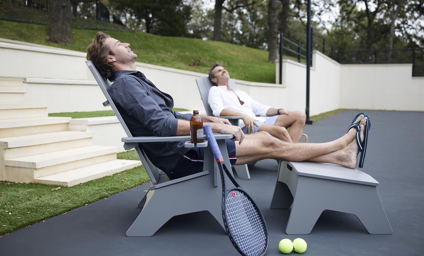 Two men relax on Ledge Lounger patio chairs with tennis rackets and balls nearby. One sips a drink as they both bask in the sun, eyes closed. Mainstay Adirondack Ottomans and steps leading to a grassy area add durability and style to the scene.