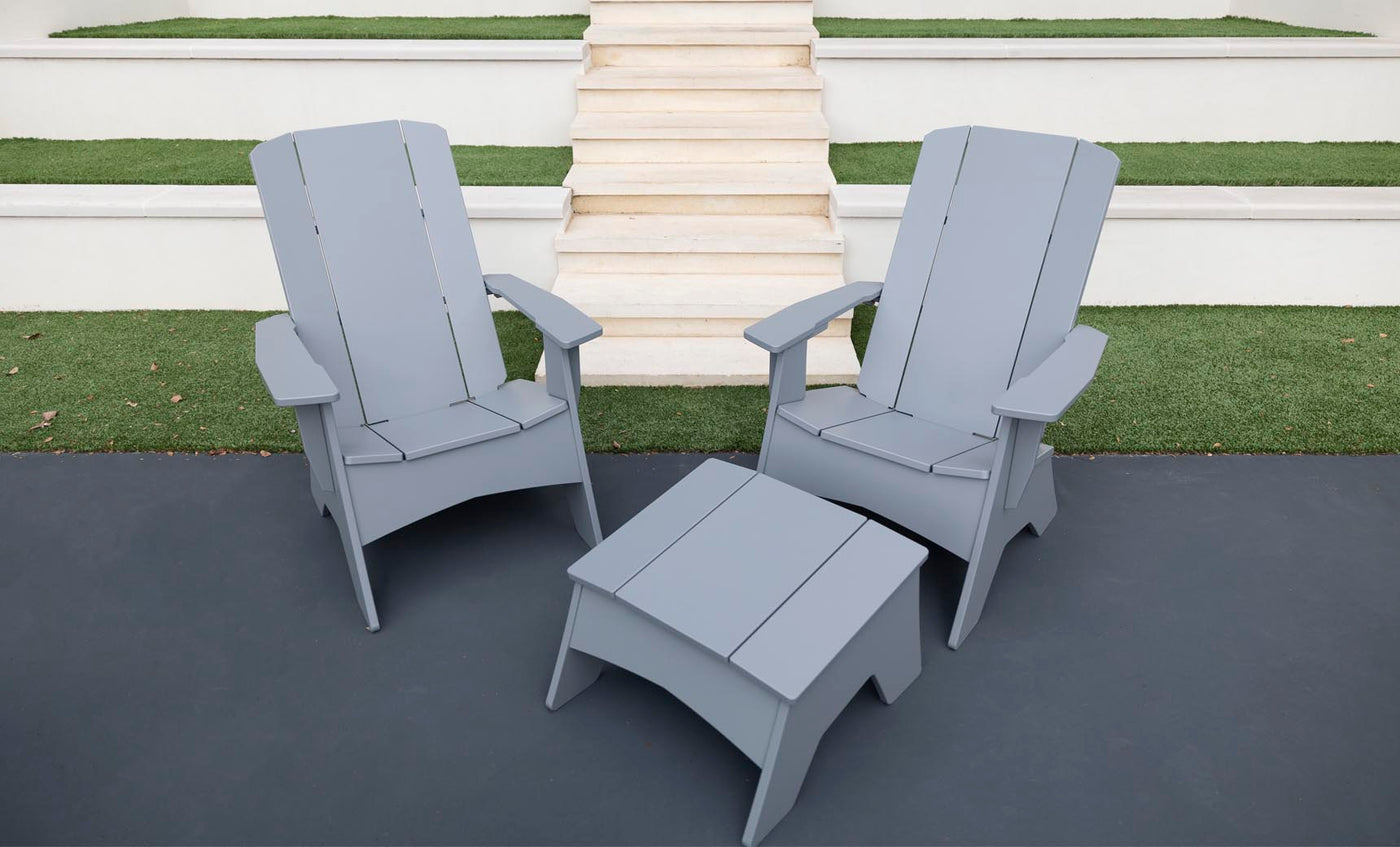 Two gray Ledge Lounger Adirondack chairs on a dark patio, showcasing durability and style, with a Mainstay Adirondack Ottoman in between. Background steps and grass patches complete the cozy outdoor setting.