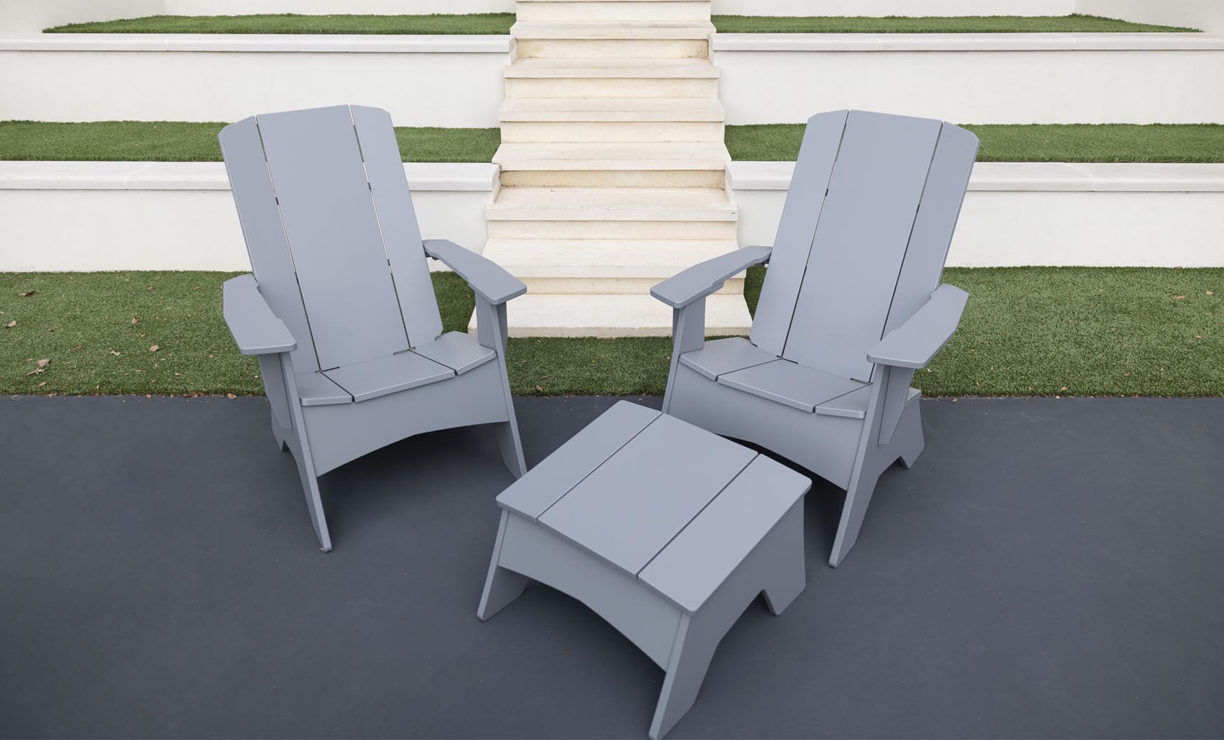 Two gray Ledge Lounger Adirondack chairs on a dark patio, showcasing durability and style, with a Mainstay Adirondack Ottoman in between. Background steps and grass patches complete the cozy outdoor setting.