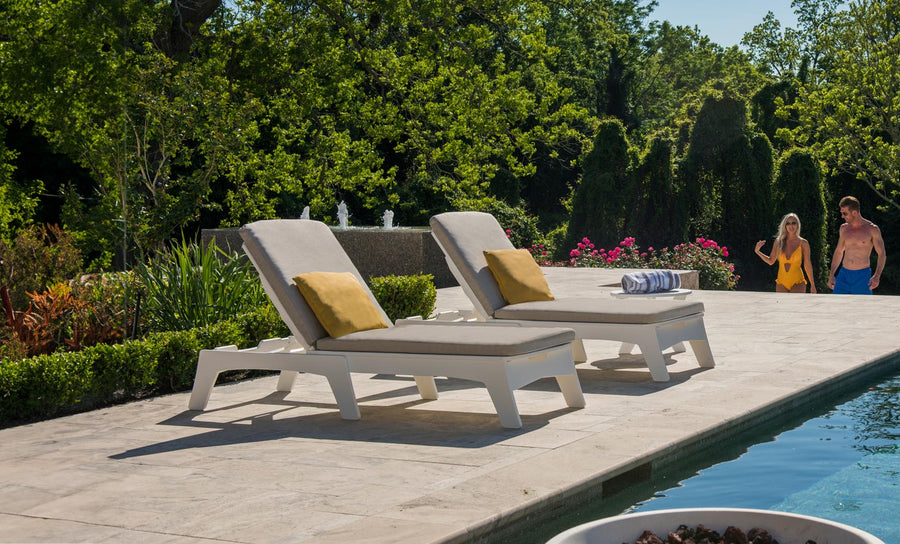 Two Ledge Lounger Mainstay Chaise lounge chairs with yellow cushions grace the stone deck. A couple in swimwear strolls by, while lush greenery enhances the serene ambiance. The pool edge glimmers invitingly in the foreground.