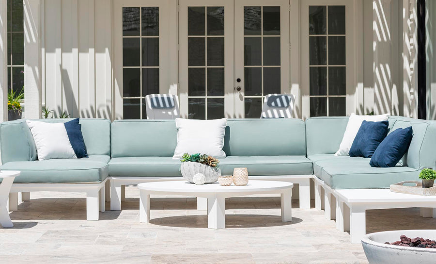 A modern patio features a minimalistic light blue sectional with white and dark blue pillows. The Mainstay Round Coffee Table by Ledge Lounger displays decor, as sunlight dances on the beige stone floor. Multi-pane glass doors complete the scene.