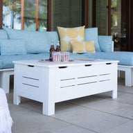 A stylish outdoor space features a blue cushioned sofa, colorful pillows, and the Mainstay Storage Coffee Table from Ledge Lounger that holds drinks. A straw bag with a white towel is on the tiled floor. Surrounded by walls with large windows, this area provides both elegance and ample storage for essentials.