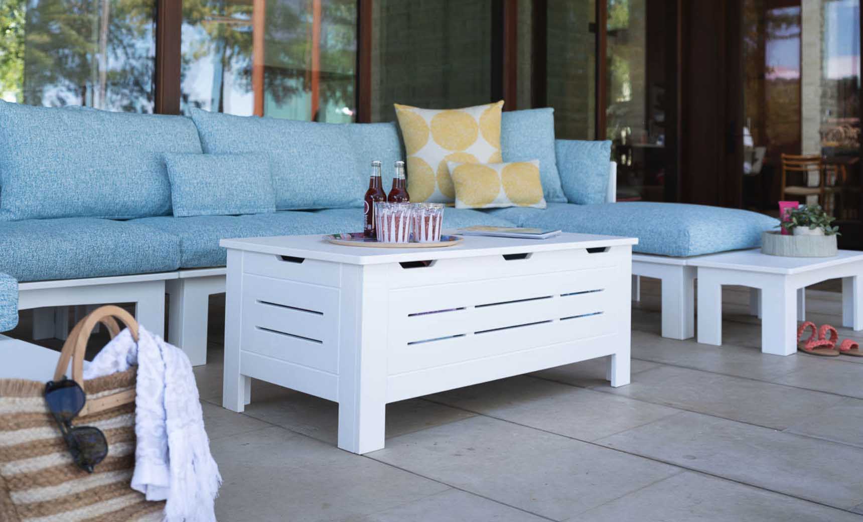 A stylish outdoor space features a blue cushioned sofa, colorful pillows, and the Mainstay Storage Coffee Table from Ledge Lounger that holds drinks. A straw bag with a white towel is on the tiled floor. Surrounded by walls with large windows, this area provides both elegance and ample storage for essentials.