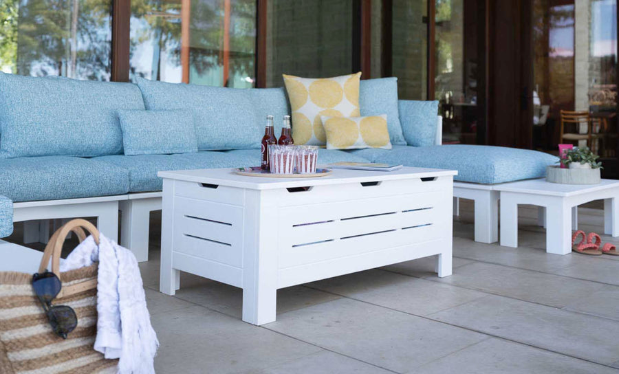 A stylish outdoor space features a blue cushioned sofa, colorful pillows, and the Mainstay Storage Coffee Table from Ledge Lounger that holds drinks. A straw bag with a white towel is on the tiled floor. Surrounded by walls with large windows, this area provides both elegance and ample storage for essentials.