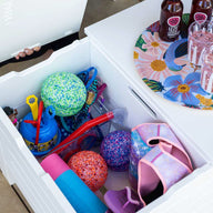 A white outdoor storage box is open, displaying colorful pool toys like balls and floats in the vibrant living area. Nearby, a Mainstay Storage Coffee Table by Ledge Lounger holds a floral tray with bottles and glasses. A person is partially visible, seated barefoot.