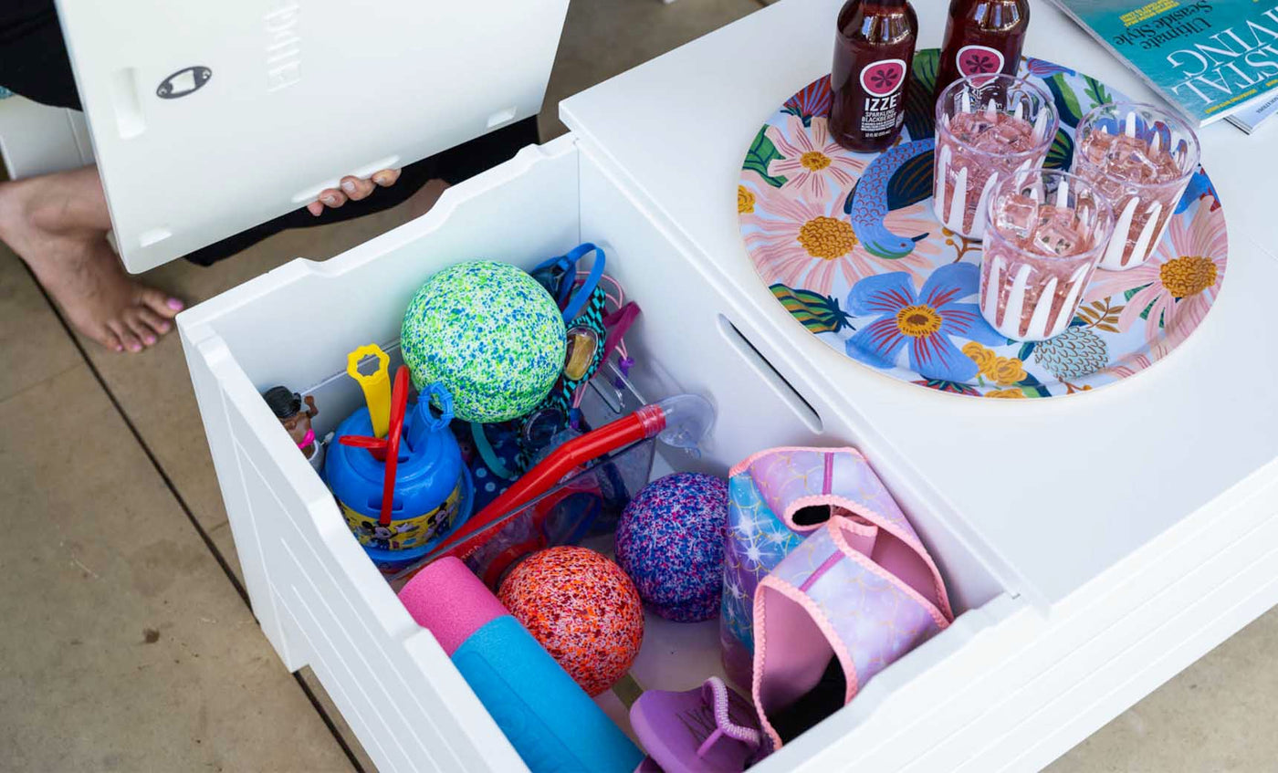 A white outdoor storage box is open, displaying colorful pool toys like balls and floats in the vibrant living area. Nearby, a Mainstay Storage Coffee Table by Ledge Lounger holds a floral tray with bottles and glasses. A person is partially visible, seated barefoot.