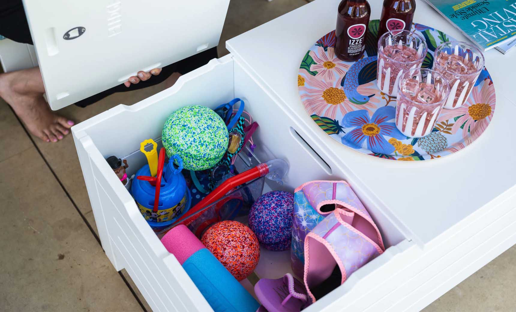 A white outdoor storage box is open, displaying colorful pool toys like balls and floats in the vibrant living area. Nearby, a Mainstay Storage Coffee Table by Ledge Lounger holds a floral tray with bottles and glasses. A person is partially visible, seated barefoot.