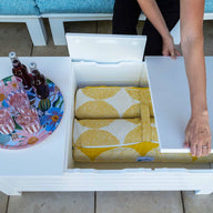 In an outdoor living space, a Mainstay Storage Coffee Table by Ledge Lounger with a white lid stores yellow-patterned blankets and supports a tray with three soda bottles and two glasses. Blue cushioned seating enhances the comfort of this inviting setting.