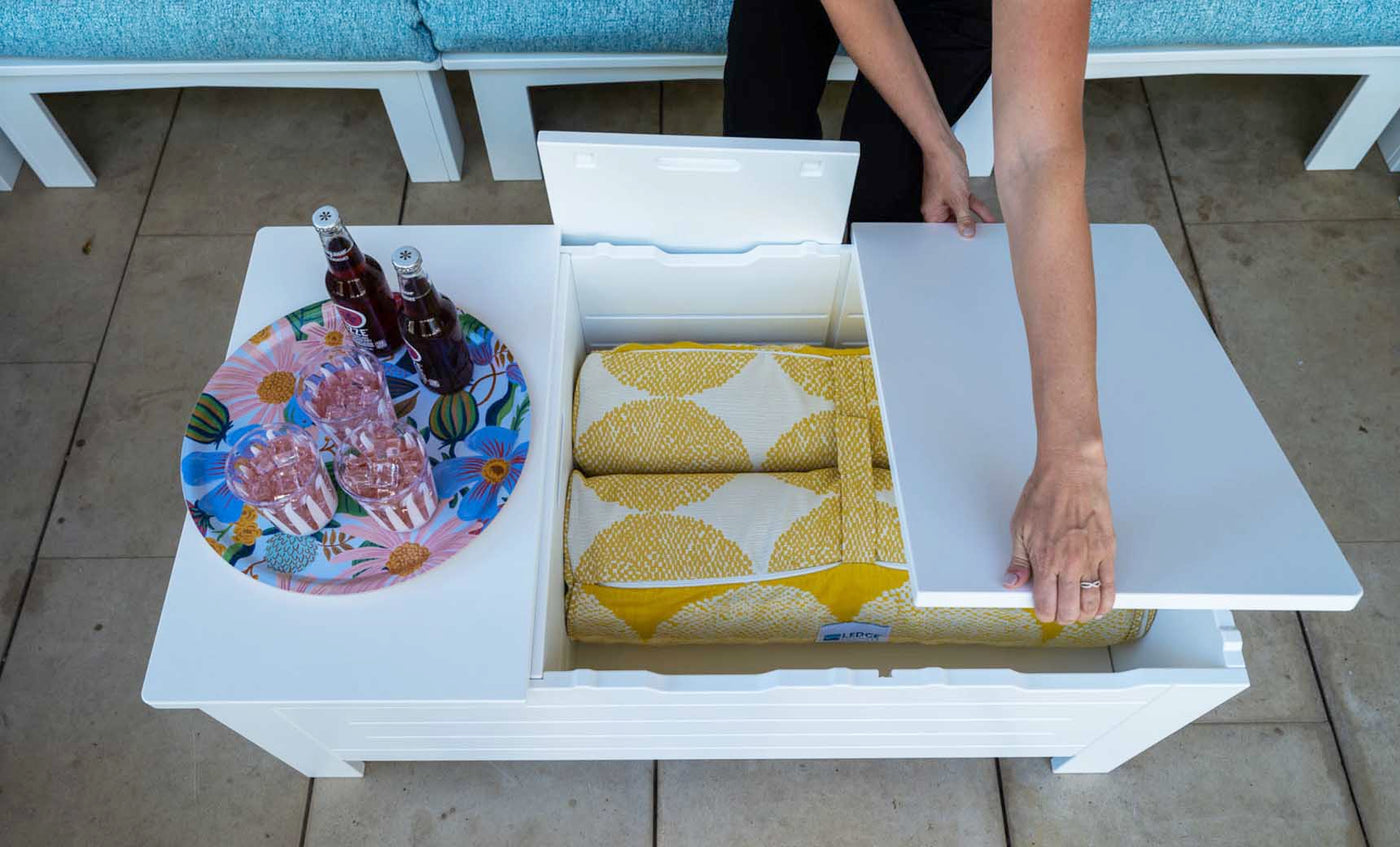 In an outdoor living space, a Mainstay Storage Coffee Table by Ledge Lounger with a white lid stores yellow-patterned blankets and supports a tray with three soda bottles and two glasses. Blue cushioned seating enhances the comfort of this inviting setting.