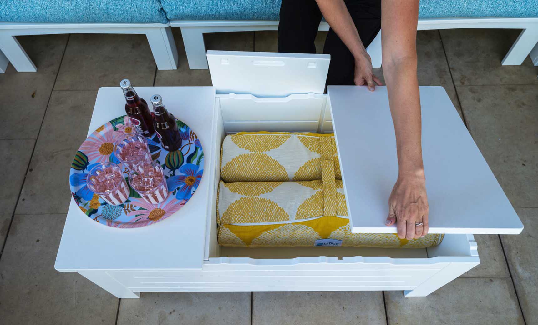 In an outdoor living space, a Mainstay Storage Coffee Table by Ledge Lounger with a white lid stores yellow-patterned blankets and supports a tray with three soda bottles and two glasses. Blue cushioned seating enhances the comfort of this inviting setting.