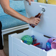 Someone is using the built-in opener on a Mainstay Storage Coffee Table (by Ledge Lounger) to open a bottle. This white coffee table, filled with colorful balls and toys, sits indoors against blue seating and yellow cushions, offering stylish storage for the living area.