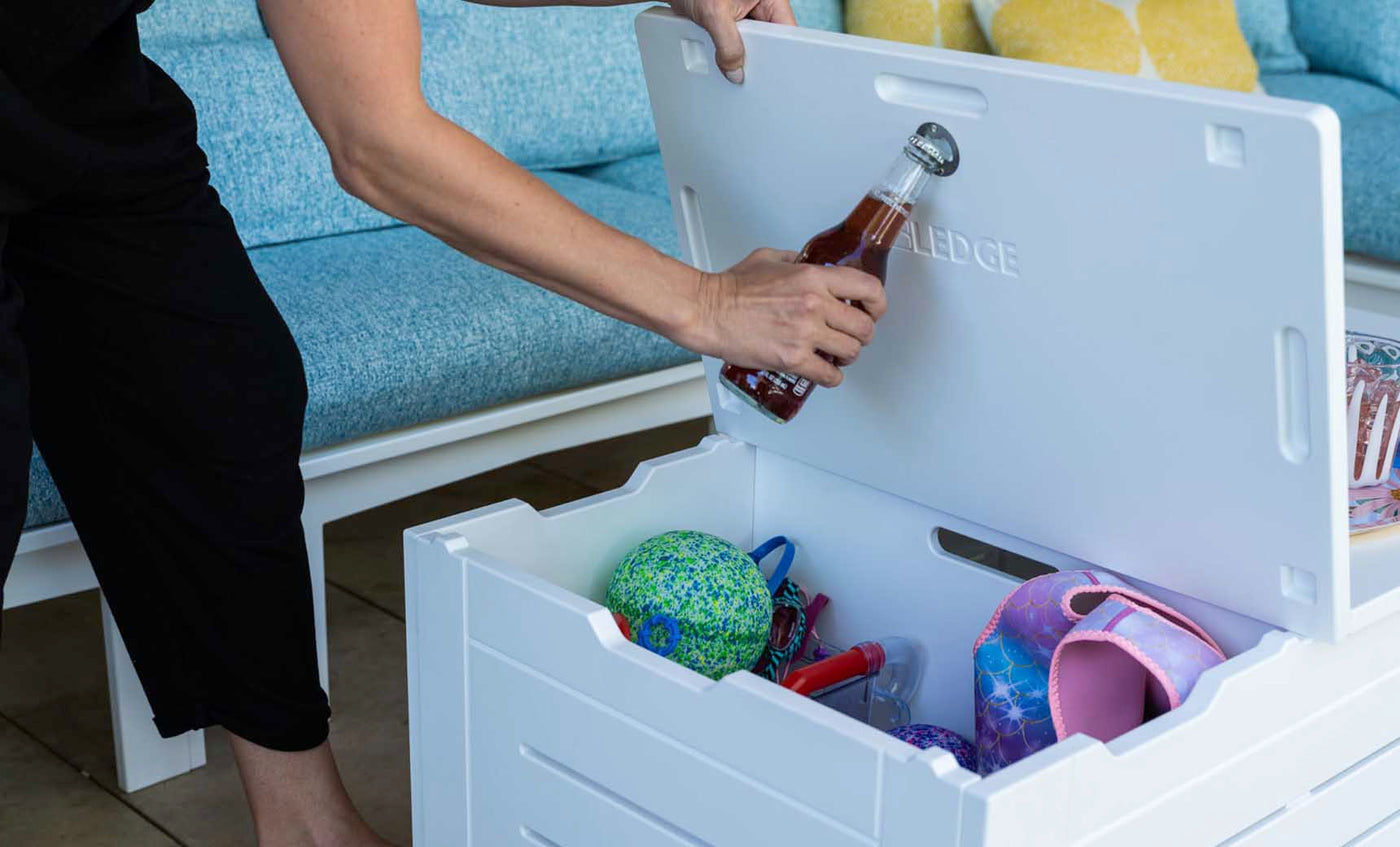 Someone is using the built-in opener on a Mainstay Storage Coffee Table (by Ledge Lounger) to open a bottle. This white coffee table, filled with colorful balls and toys, sits indoors against blue seating and yellow cushions, offering stylish storage for the living area.