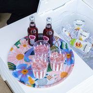 A Ledge Lounger floral tray with three drinks and glasses sits on the Mainstay Storage Coffee Table, set in an inviting outdoor space. Nearby, a bucket with ice holds bottled drinks, while a partially visible person sits on the left and a magazine rests on the table.
