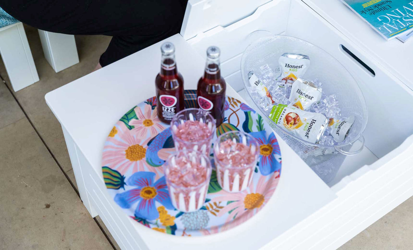 A Ledge Lounger floral tray with three drinks and glasses sits on the Mainstay Storage Coffee Table, set in an inviting outdoor space. Nearby, a bucket with ice holds bottled drinks, while a partially visible person sits on the left and a magazine rests on the table.
