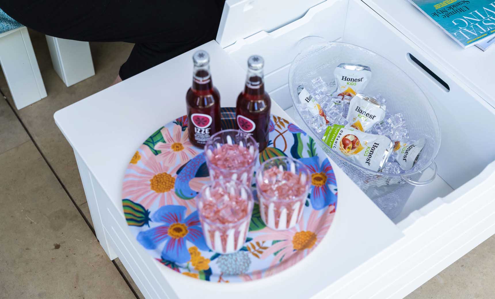 A Ledge Lounger floral tray with three drinks and glasses sits on the Mainstay Storage Coffee Table, set in an inviting outdoor space. Nearby, a bucket with ice holds bottled drinks, while a partially visible person sits on the left and a magazine rests on the table.