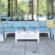 Outdoor living space with a light blue sectional sofa and yellow cushions. A Mainstay Storage Coffee Table by Ledge Lounger is centered, displaying bottles and a magazine. A straw hat, striped tote, and potted plants rest nearby. Large windows reflect the surrounding greenery.