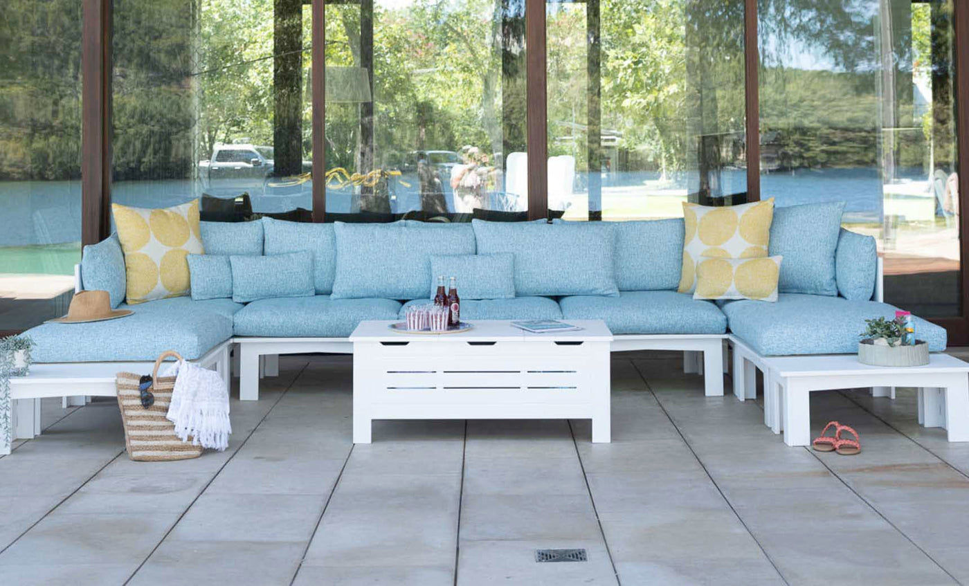 Outdoor living space with a light blue sectional sofa and yellow cushions. A Mainstay Storage Coffee Table by Ledge Lounger is centered, displaying bottles and a magazine. A straw hat, striped tote, and potted plants rest nearby. Large windows reflect the surrounding greenery.