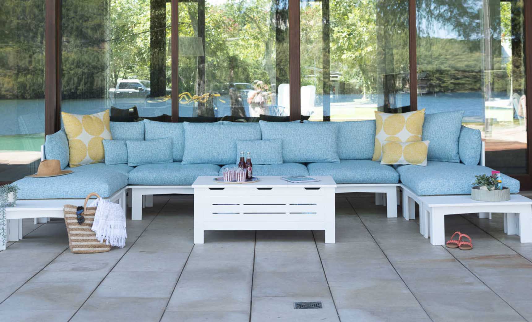 Outdoor living space with a light blue sectional sofa and yellow cushions. A Mainstay Storage Coffee Table by Ledge Lounger is centered, displaying bottles and a magazine. A straw hat, striped tote, and potted plants rest nearby. Large windows reflect the surrounding greenery.