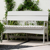 A white Mainstay Bench from Ledge Lounger, featuring a patterned cushion, is set on a stone patio with lush greenery and a vine-covered white wall. Pink flowers bloom on the left, enhancing this stylish, all-weather outdoor retreat.