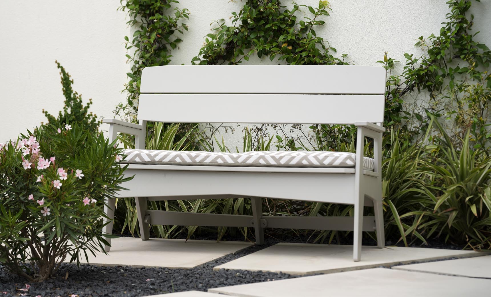 A white Mainstay Bench from Ledge Lounger, featuring a patterned cushion, is set on a stone patio with lush greenery and a vine-covered white wall. Pink flowers bloom on the left, enhancing this stylish, all-weather outdoor retreat.