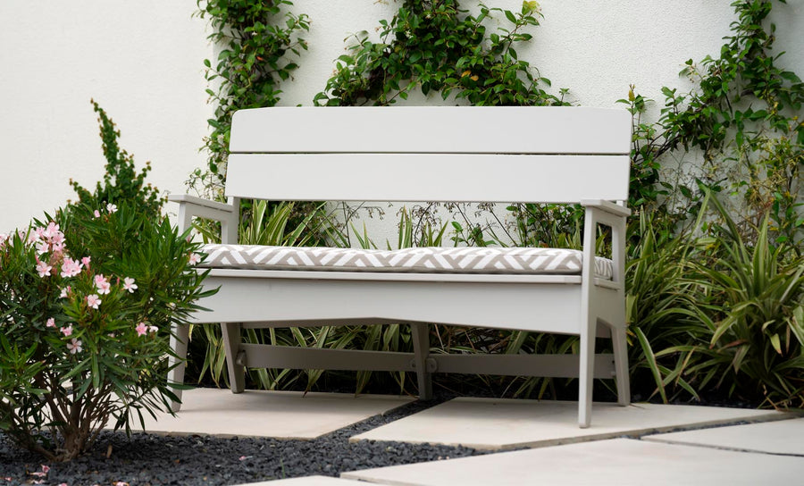 A white Mainstay Bench from Ledge Lounger, featuring a patterned cushion, is set on a stone patio with lush greenery and a vine-covered white wall. Pink flowers bloom on the left, enhancing this stylish, all-weather outdoor retreat.