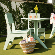 Enjoy a blissful outdoor dining setup with Ledge Lounger’s Mainstay Dining Side Chairs and two light green tables on artificial grass. A woven basket with a magazine and pink water bottle sits below, while a pink cloth drapes over one chair. Yellow flowers add charm to this high-quality scene.